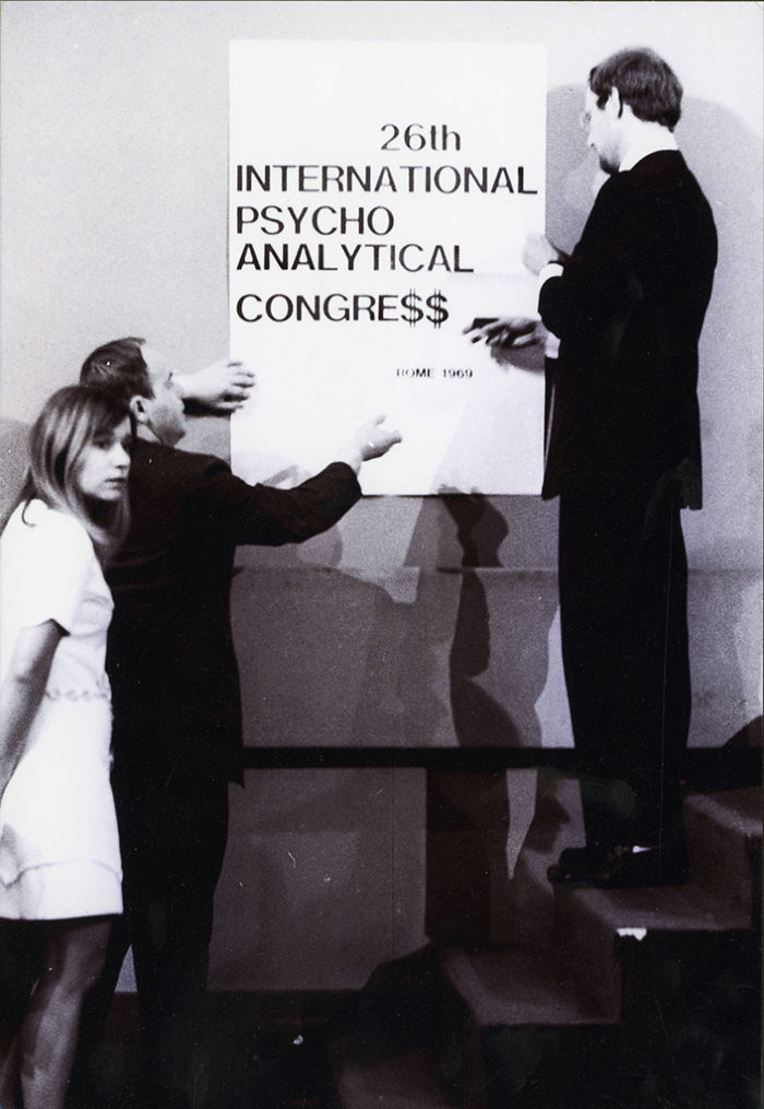 Elvio Fachinelli con Berthold Rothschild e Marianna Bolko durante il "controcongresso", accanto a un cartello con la scritta "26th International Psycho Analytical Congre$$” (© Lisetta Carmi 1969, Collezione privata)