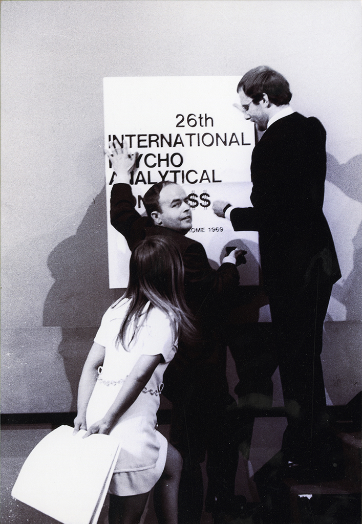 Elvio Fachinelli con Berthold Rothschild e Marianna Bolko durante il "controcongresso", accanto a un cartello con la scritta "26th International Psycho Analytical Congre$$” (© Lisetta Carmi 1969, Collezione privata)